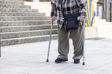 An elderly individual is seen from the waist down utilizing two walking canes for mobility assistance on a city street