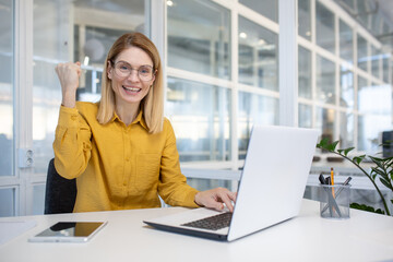 Excited businesswoman celebrating success while working on a laptop in a modern office setting, showcasing business and achievement concepts.
