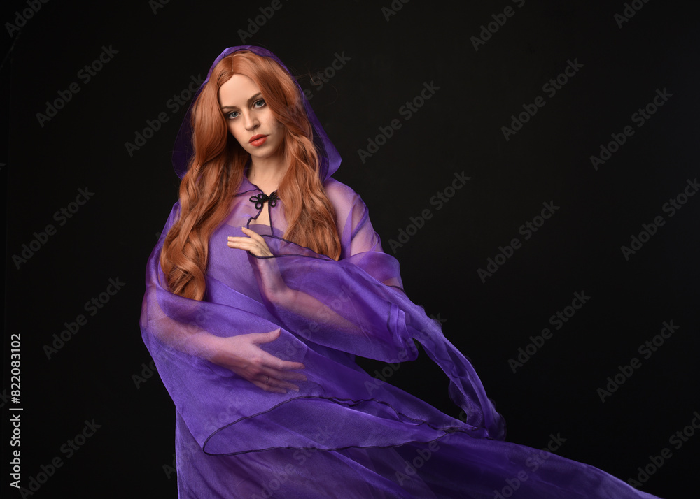 Wall mural close up portrait of beautiful female model with long red hair, wearing  purple fairy fantasy cloak gown. Elegant gestural hand poses casting magic spell. Isolated on dark black studio background