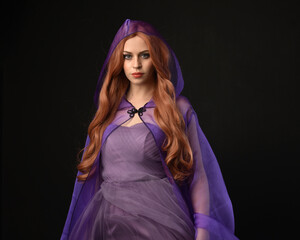 close up portrait of beautiful female model with long red hair, wearing  purple fairy fantasy cloak gown. Elegant gestural hand poses casting magic spell. Isolated on dark black studio background