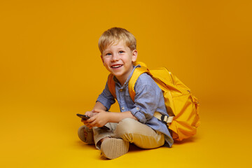 Positive schoolboy with yellow rucksack sitting on studio floor with crossed legs, using mobile phone and looking at camera excited. Isolated over yellow background.