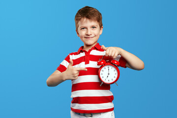Delighted little boy in casual red striped shirt, pointing index finger on red alarm clock. Posing...