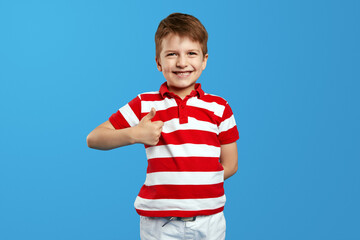 Little preschool boy smiling and wearing red striped polo shirt, posing on blue background while...