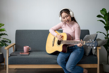A woman is playing the guitar on a couch. She is wearing headphones and smiling. The couch is...