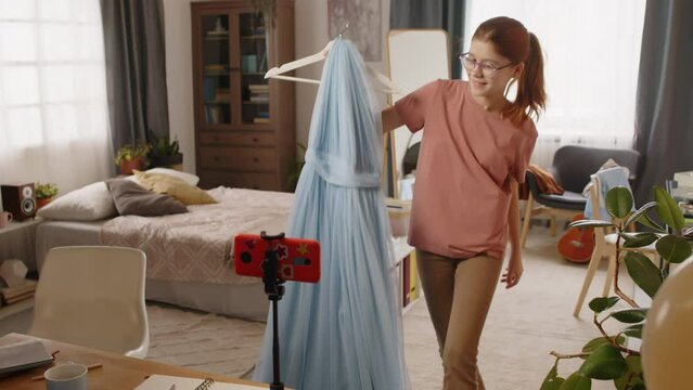 Cute teenage girl with blue evening dress moving towards smartphone on desk, pressing record button and presenting her new gown bought for prom party to online audience