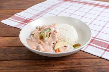 A savory meal presented with creamy chicken and vegetables next to a scoop of white rice on a wooden surface with a checkered napkin