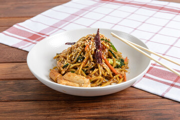 A plate of stir-fried noodles mixed with chicken, vegetables, and topped with sesame seeds, served on a wooden table with chopsticks