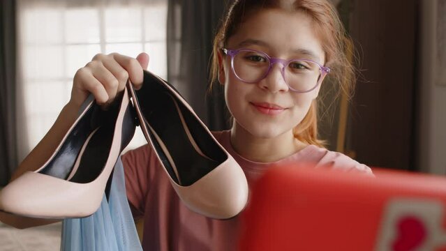 Cute smiling teenage girl in eyeglasses looking at smartphone camera and boasting with her new shoes and evening gown bough for prom celebration to online audience