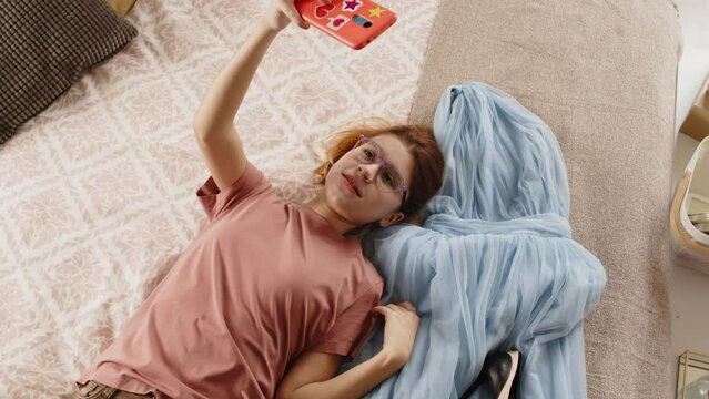High angle of teenage girl with smartphone lying on double bed next to new evening own and pair of shoes on high heels and speaking to online audience or recording video before prom party
