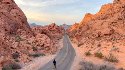 A lone woman walks down the center of an empty desert road in a beautiful canyon surrounded by pastel red rock at sunrise - Valley of Fire State Park, Nevada, USA