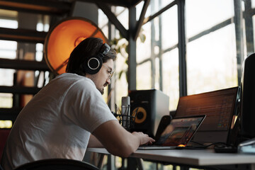 Man working on an audio project in a home studio, wearing headphones and using multiple screens....