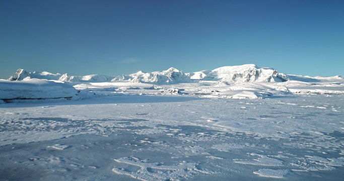 Fly over snowy mountain landscape in Antarctica. Polar frozen ocean landscape covered by snow under blue sunny sky. Discover the beauty of South Pole. Antarctica travel and exploration background