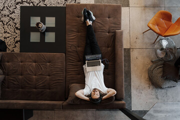 Top view of a man relaxing on a brown couch with a laptop on his chest in a modern living room.