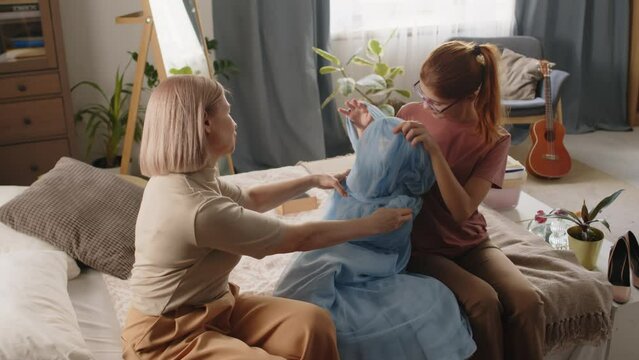Cute adolescent daughter and her mother in casualwear sitting on bed in domestic room and discussing new fatin evening gown before celebration of prom in highschool