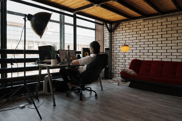 Man working on computer in a modern industrial loft office with large windows, minimalist furniture, and comfortable ambiance.