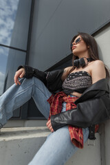 fashionable beautiful urban woman with sunglasses in stylish denim casual clothes sits in the city near a building