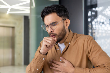 Young man coughing in modern office environment, hand on chest