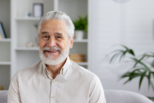 Close-up portrait of a smiling gray-haired senior man at home, sitting on a sofa in a white shirt and with a beard, smiling at the camera