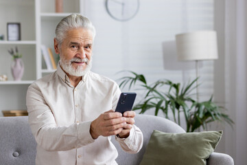 Portrait of an older gray-haired man in a white shirt sitting on the couch at home, holding a mobile phone and smiling at the camera