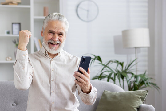 Happy gray-haired senior man is at home, sitting on the couch, looking at the screen of the phone he is holding and rejoicing in success, showing a victory gesture with his hand - Powered by Adobe