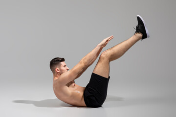 A shirtless man demonstrates the hollow body hold exercise while wearing black shorts and sneakers....