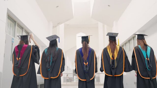 Five women in graduation gowns are standing in a hallway. They are all holding their graduation caps and gowns