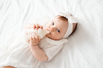 Newborn baby girl sucks a bottle of milk in white clothes on the bed at home on her back, cute baby...