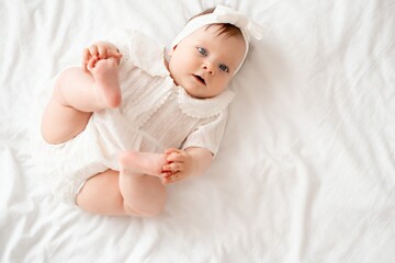 Newborn baby girl plays with her legs pushing them into her mouth in white clothes on the bed at...