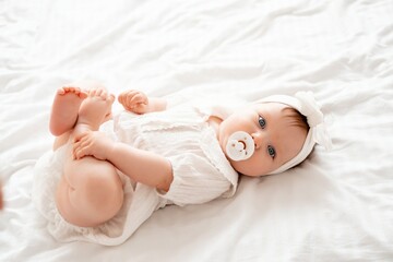 Cute newborn baby girl sucks a pacifier in white clothes on the bed at home on her back, cute baby...