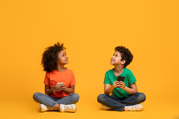 Two African American children sit on the floor, engrossed in their smartphones. Their attention is...
