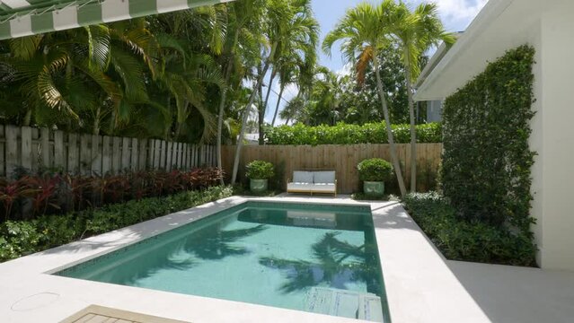Outdoors residential area featuring small rectangular swimming area. Spacious pool surrounded by patio with white tiles and striped awning. Lush greenery and tall hedges