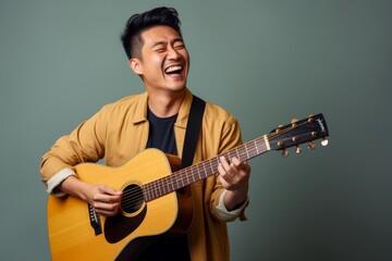 Portrait of a cheerful asian man in his 30s playing the guitar while standing against blank studio...