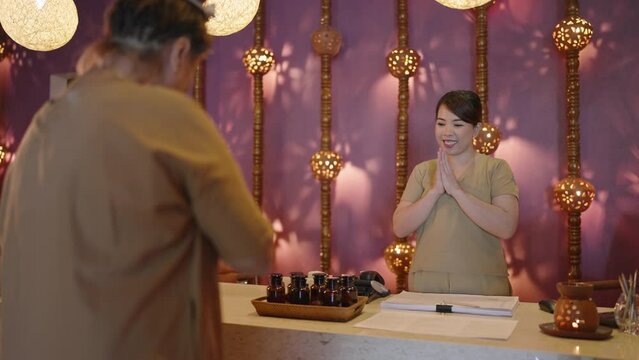 Senior asian female client chooses care procedures smells aroma oils at authentic spa center. Elderly multiracial woman talks to receptionist at atmospheric retreat center, picks treatment programs
