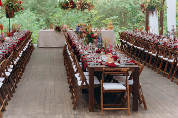A large, long, wooden table and chairs, with dishes, flowers, candles. Pavilion in nature. Wedding banquet.