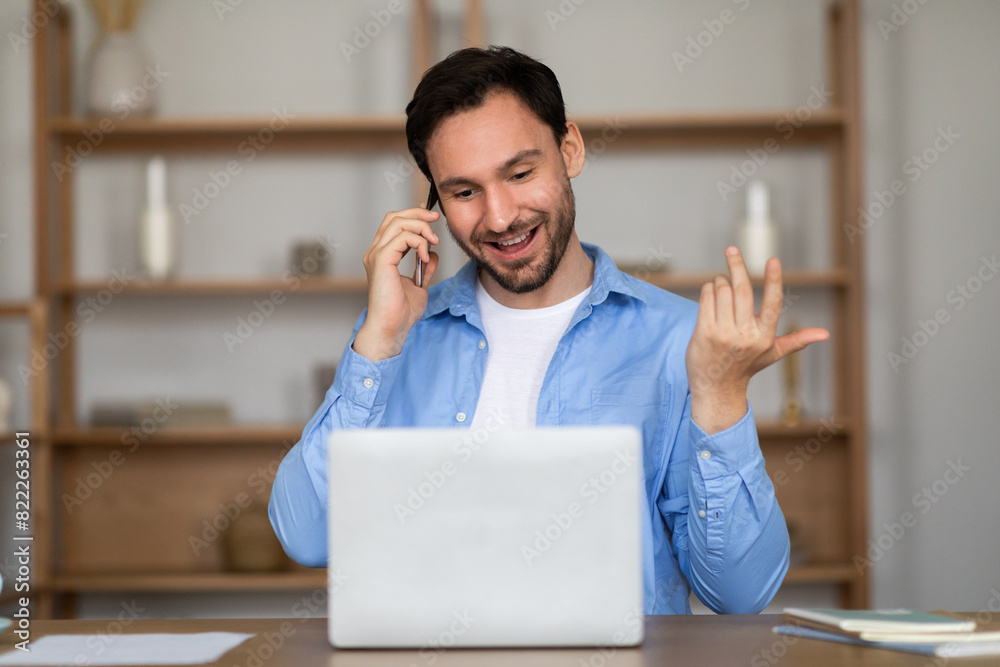 Poster a man is seated in front of a laptop, engaged in a phone conversation. he appears focused on the scr