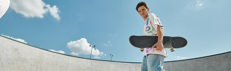 A young man holds a skateboard, standing in a vibrant skate park on a sunny day, exuding a sense of...