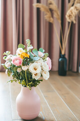 Vibrant assorted flowers fill a textured pink vase, resting on a wooden floor against elegant pink curtains