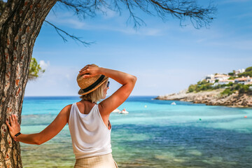 A woman in a straw hat standing by the shore under a pine tree, gazing at the clear blue sea in...