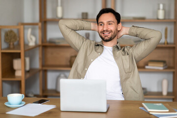 A young man with a cheerful expression sits back at his desk in a well-lit, contemporary office...