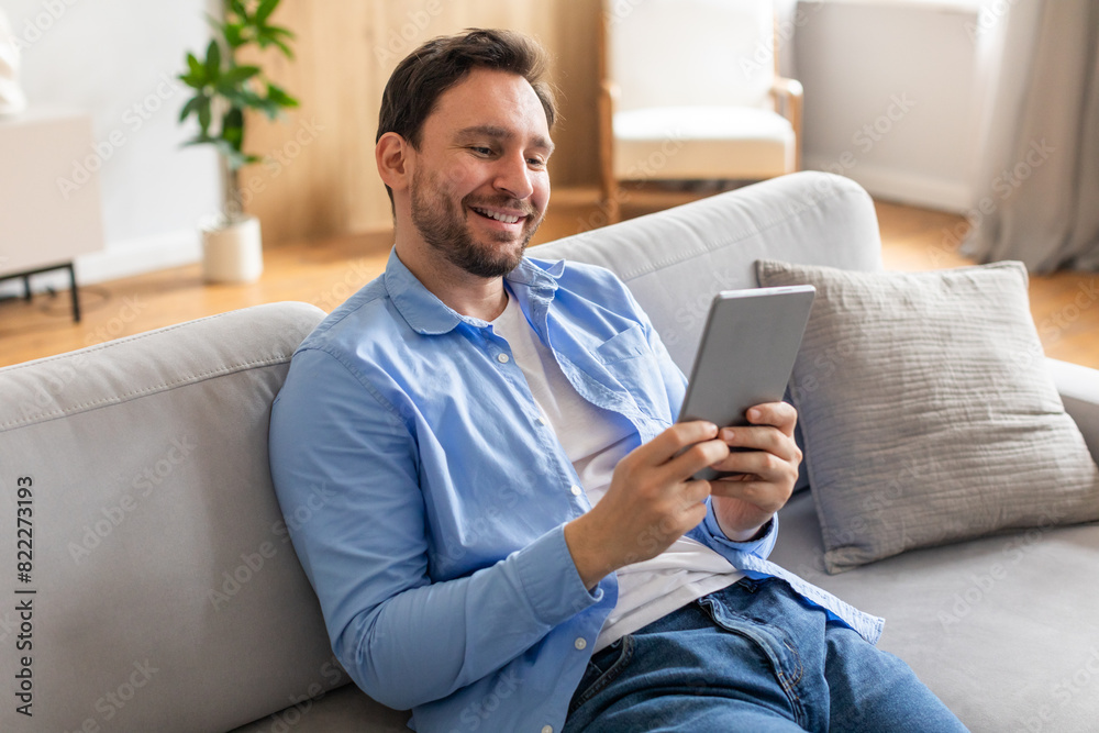 Poster A man is seated on a couch, engrossed in using a tablet computer. He is focused on the screen, tapping and scrolling through content. The room is well-lit