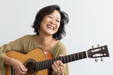 Portrait of a happy asian woman in her 50s playing the guitar isolated in light wood minimalistic...