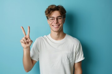 Fototapeta premium Blue background, portrait, guy and peace fingers in studio for hope, self-love, and icon. Face of joyful Asian guy model, v sign, hands for triumph, good mood, and freedom emoji.
