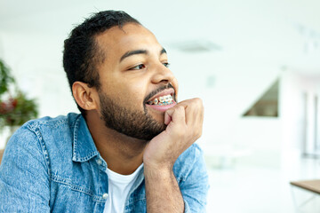 happy african american man with braces smiles and looks away, man in denim shirt takes care of teeth