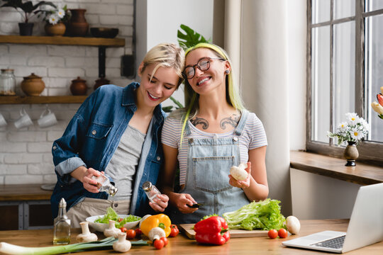 Two affectionate caucasian young lesbians girlfriends lgbtq couple cooking, preparing food at home kitchen together, cuddling and hugging, watching culinary recipe show at home kitchen - Powered by Adobe
