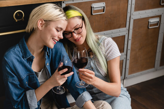 Top view on young diverse caucasian lgbtq lesbian couple hugging kissing cuddling drinking wine together sitting at floor home kitchen, looking at each other, having romantic date.