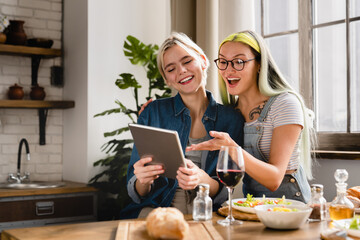 Passionate romantic lgbtq lesbian couple using digital tablet for social media, e-shopping, buying presents online, remote work while having breakfast together at home kitchen