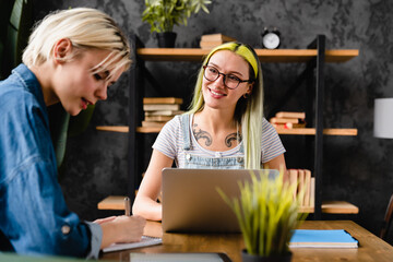 Two young coworkers female lgbtq couple girlfriends colleagues working together at coworking area,...