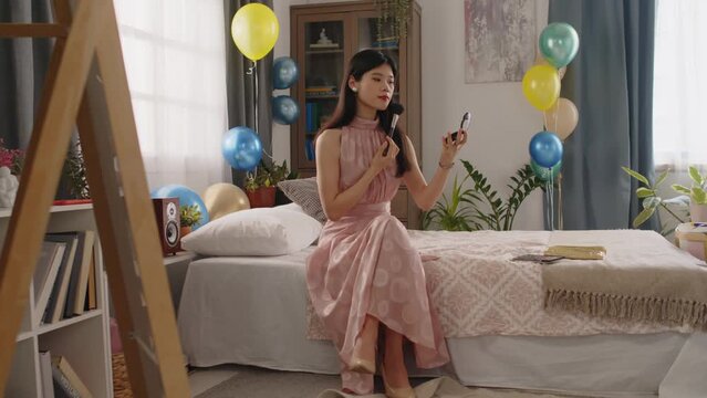 Youthful Asian girl in pink evening dress sitting on double bed in domestic room and looking in small mirror while applying powder on face before going to prom party