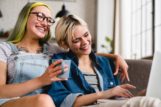 Closeup happy cheerful young sisters girlfriends lgbtq lesbian couple chilling on the sofa, hugging cuddling and watching movie series film webinar on laptop together at home online
