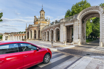A cobbled road with a red vehicle on the zebra crossing passing beneath an old building with arches and passages at the royal site of Aranjuez, Madrid